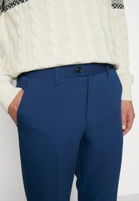 Navy blue tailored trousers with smooth texture, side pockets, and a black button closure, paired with a cream cable-knit sweater.