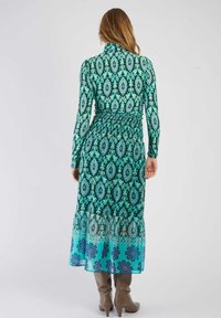 Robe maxi à manches longues en turquoise avec des motifs paisley et floraux complexes, dotée d'une taille smockée et d'un ourlet dégradé. Associée à des bottes beiges jusqu'aux genoux.