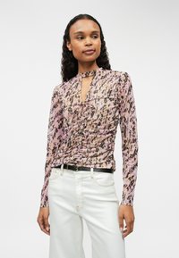 Bluzka z długim rękawem w różowo-brązowy wzór zwierzęcy, z dekoltem w kształcie łezki i marszczeniami, połączona z białymi spodniami o wysokim stanie.