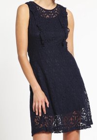 Navy blue lace dress with a sleeveless design, featuring ruffled accents along the neckline and a floral pattern, finished with a scalloped hem.