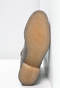 Hnědá podrážka boty z texturovaného gumového materiálu, s diagonálními drážkami pro lepší grip a hladkým, mírně vyvýšeným okrajem kolem obvodu.