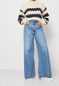 Henkilö, jolla on päällä vaaleanbeige ja tummansiniraidallinen paksu neule, korkeavyötäröiset leveälahkeiset vaaleansiniset farkut ja valkoiset lenkkarit, kädet taskuissa.