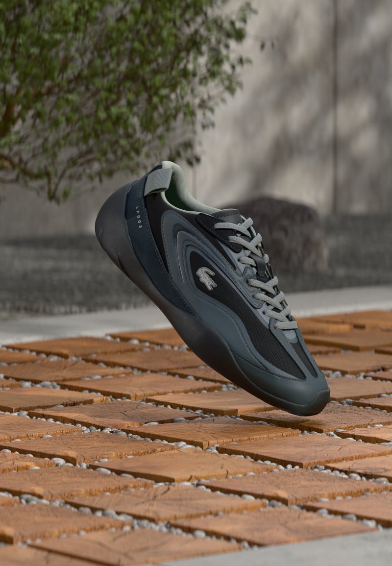 Baskets Lacoste en gris foncé et noir avec une tige en tissu lisse, une semelle en caoutchouc texturée, et des lacets gris clair ; dotées d'une patte de tirage arrière.