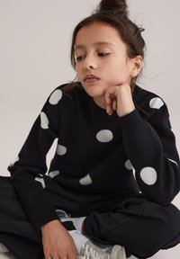 Pull noir à gros pois blancs, col rond, poignets côtelés, associé à un pantalon noir et des baskets argentées. Texture tricotée confortable.