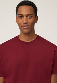 T-shirt di cotone color bordeaux con collo tondo, maniche corte e un piccolo logo di cane ricamato sul lato sinistro del petto. Tessuto morbido.