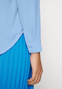 Camicia azzurro chiaro a maniche lunghe e texture con polsini buttonati, abbinata a una gonna blu brillante a pieghe e fluida.