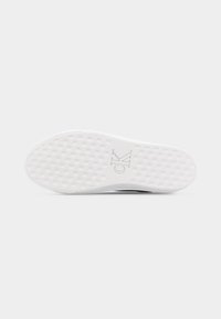 Λευκή σόλα αθλητικού παπουτσιού με σχέδιο ελέγχου στην πέλμα και ανάγλυφο λογότυπο «cK» στο κέντρο σε ανοιχτό γκρι φόντο.