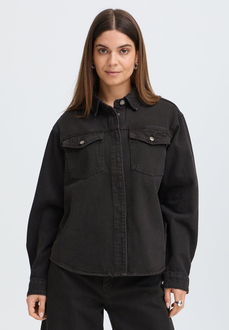 Femme aux cheveux bruns et raides portant une veste en jean noire avec des poches poitrine boutonnées, debout devant un fond clair uni.