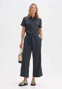 Dunkelgrüner Overall mit kurzen Ärmeln, Knopfverschluss vorne, zwei Brusttaschen, gebundenem Taillengürtel, weit geschnittenen Beinen, kombiniert mit grünen Sandalen und einer geflochtenen Tasche.