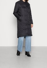 Abrigo largo acolchado negro con cuello alto, botones a presión y dos bolsillos laterales, combinado con vaqueros de pierna ancha en azul claro y botas negras de tacón.