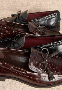 Două perechi de mocasini: una neagră, cealaltă maro închis cu un model texturat de crocodil. Ambele au fundațe decorative din piele și orificii argintii.