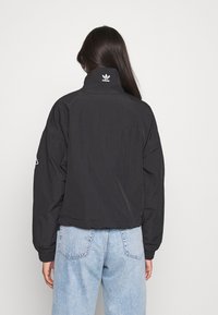 Svart vindjacka med hög krage och en vit logotyp på baksidan. Har sänkta axlar och elastiska muddar. Matcher med ljusblåa jeans.