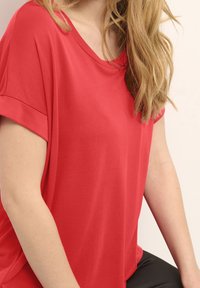 Top rosso a maniche corte realizzato in tessuto morbido, con vestibilità relax e scollatura ampia, abbinato a pantaloni scuri. Tessuto liscio senza fantasie.