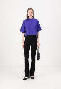 Maglia cropped oversize viola con maniche corte abbinata a pantaloni neri a campana e scarpe nere, mentre tiene una piccola borsa nera.
