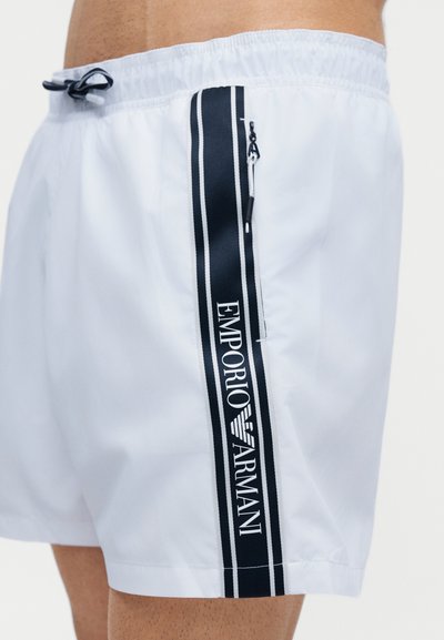 Λευκά μαγιό σορτς με μαύρη κάθετη ρίγα και τσεπάκι με φερμουάρ που φέρει το λογότυπο "Emporio Armani" στη μέση και το πάνω μέρος του μηρού ενός ανδρικού μοντέλου.