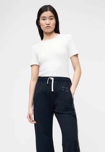 Νεαρή γυναίκα με μακριά μαύρα μαλλιά, ντυμένη με λευκή κοντομάνικη μπλούζα και μπλε σκούρο παντελόνι με φαρδιά πόδια, συρόμενο λευκό κορδόνι και λογότυπο.