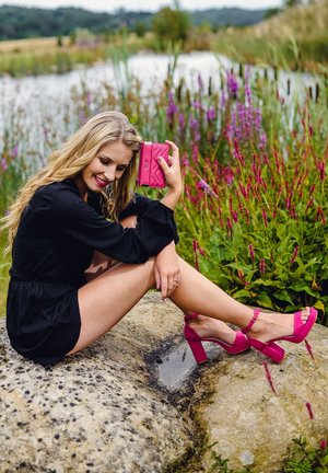 Pink quilted wallet and matching high-heeled sandals, worn with a black dress, seated on a stone near a pond and flowers.