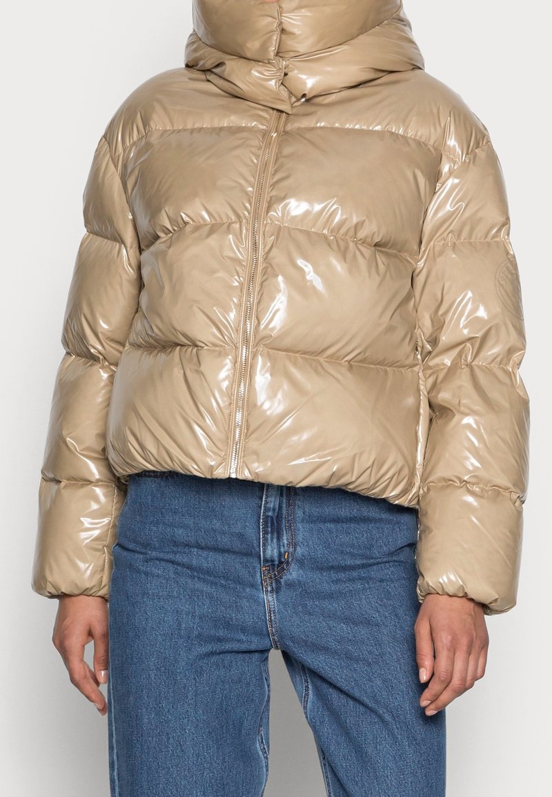 Beżowa, błyszcząca kurtka puchowa z wysokim kołnierzem, przednim zamkiem i poziomym pikowaniem. Połączona z niebieskimi dżinsami z wysokim stanem.