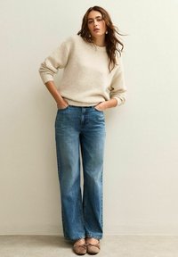 Suéter texturizado de color crema con cuello acanalado combinado con jeans de mezclilla azul de tiro alto y corte suelto. La persona está de pie de manera casual.