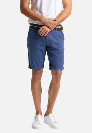 Uomo in piedi, indossa una t-shirt bianca, pantaloncini blu con risvolto e cintura a righe, e sneakers bianche, con le mani intrecciate davanti.