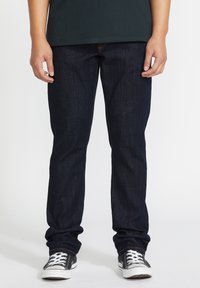 Jean en denim bleu foncé avec un design à jambe droite, mettant en avant des coutures visibles, associés à des baskets noir et blanc.
