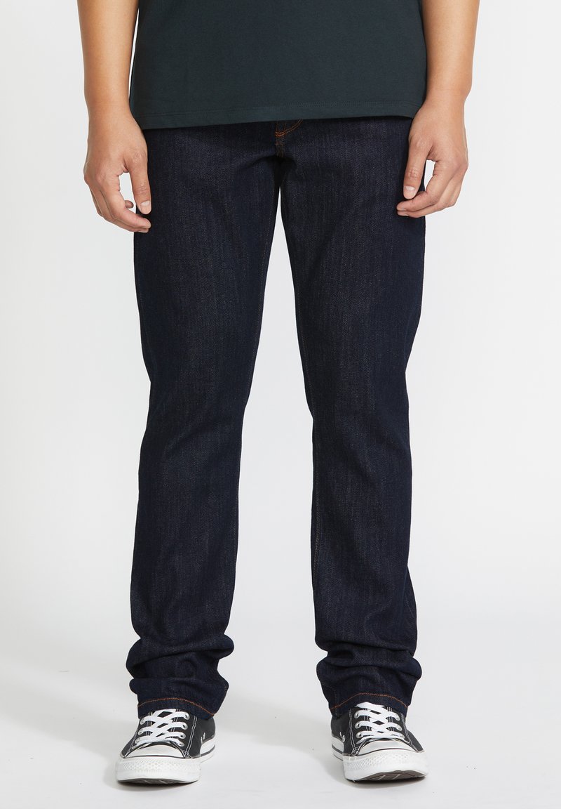 Jean en denim bleu foncé avec un design à jambe droite, mettant en avant des coutures visibles, associés à des baskets noir et blanc.