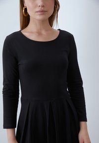 Black long-sleeve dress with a scoop neckline and pleated skirt detail, made from a soft, stretchy fabric. Gold hoop earrings visible.