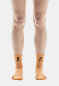 Oranje enkelsokken met een zwart logo aan de voorkant, ribbelboord, naadloze teen en zachte textuur, zichtbaar gedragen op blote voeten.
