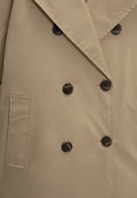 Palton bej cu un design cu dublu rând de nasturi, rever larg și nasturi maro închis. Dispune de un buzunar lateral și o textură netedă.