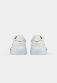 Weiße Wildledersneaker mit strukturiertem Finish, ausgestattet mit einer glatten Fersenschlaufe und schwarzen Akzenten auf der Gummisohle. Von hinten präsentiert.
