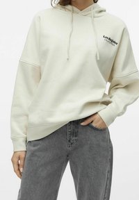 Persona che indossa una felpa color avorio con la scritta "Los Angeles California" sul petto e jeans neri scoloriti, con una mano in tasca, su sfondo bianco.