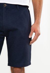 Shorts chino bleu marine en coton avec zip, fermeture à bouton, poches latérales et ourlet replié. La texture semble lisse et durable.