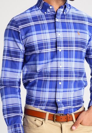 Homme portant une chemise à carreaux bleue et blanche boutonnée, rentrée dans un pantalon beige avec une ceinture en cuir marron, main dans la poche.