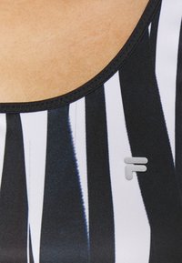 Černobílý pruhovaný sportovní top s kulatým výstřihem, který má stříbrné logo a hladkou, elastickou texturu látky.