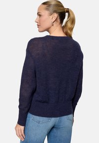 Femme avec queue de cheval blonde portant un pull douillet bleu marine et un jean bleu clair, vue de dos sur un fond blanc uni.