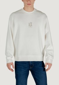 Weißer Pullover mit rundem Ausschnitt und gerippten Bündchen. Hat ein dezentes Logo in hellbeige in der Mitte. Kombiniert mit blauen Jeans.