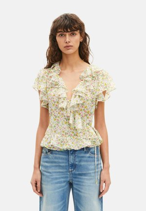 Vrouw draagt een lichte, bloemige blouse met ruches en korte mouwen, gecombineerd met lichtblauwe denim jeans, staand tegen een effen achtergrond.