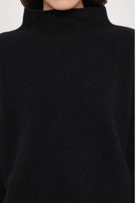 Femme portant un pull en laine noir à col montant, montrant la texture et la coupe des épaules à la partie supérieure de la poitrine.