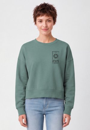 Junge Person mit kurzen braunen Haaren trägt grünes Sweatshirt mit Sonnen-Tarot-Kartendesign, kombiniert mit hellblauen Jeans, steht vor weißem Hintergrund.