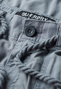 Szare szorty z materiału z plecionym sznurkiem, z ciemnym guzikiem i czarną metką, na której w białym tekście widnieje napis "Superdry".