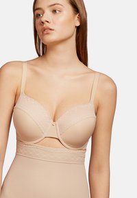 Sostén de encaje beige con ribetes, con aros, tirantes ajustables y un corte en forma de ojo de cerradura en el centro para un look sin costuras. Textura de tela suave.