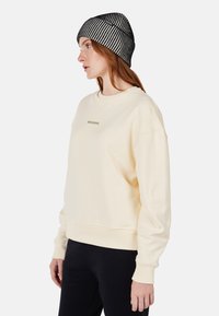Sudadera crema con puños y bajo de canalé, que presenta un pequeño logo en el pecho. Lleva puesta una boina de punto a rayas en blanco y negro.