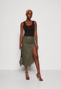 Black sleeveless bodysuit paired with a fitted olive green skirt featuring a side slit and ruched detailing, accented by strappy black heels.