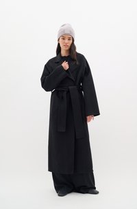 Černý vlněný trench coat s širokými rukávy, pasem na opasek a klopovým límcem. V kombinaci se světle šedou žebrovanou čepicí.