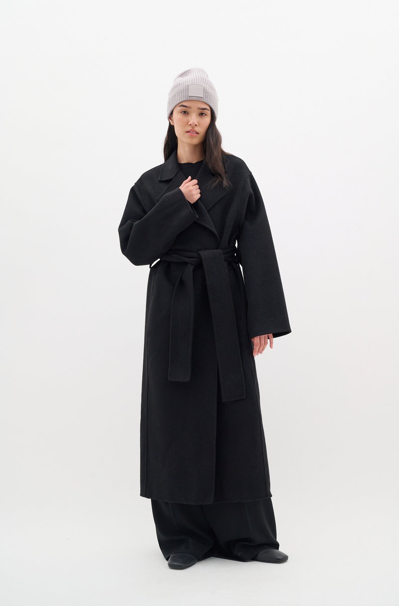 Černý vlněný trench coat s širokými rukávy, pasem na opasek a klopovým límcem. V kombinaci se světle šedou žebrovanou čepicí.