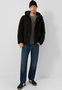 Giacca nera imbottita con cappuccio, chiusura con zip e bottoni a pressione. Indossata sopra una maglietta grigia scura e jeans in denim blu. Sneakers nere con dettagli.