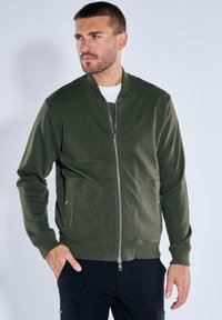 Dark green bomber jacket with a smooth texture, front zipper fastening, side pockets, and ribbed cuffs. Simple, minimal design.