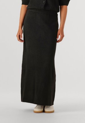 Zwarte gebreide maxi-rok met geribde tailleband en zijsplit, gecombineerd met lichtgekleurde sneakers. Zachte textuur en aansluitende silhouet.