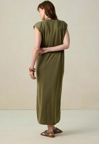 Femme de dos portant une robe maxi sans manches olive et des sandales imprimées léopard, sur un fond beige uni.