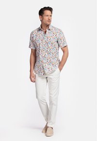 Chemise à manches courtes avec un fond blanc et des motifs abstraits colorés ; associée à un pantalon beige clair et des chaussures à enfiler beiges.